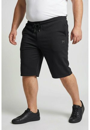 Mann trägt schwarze Shorts mit Kordelzug, weißes Hemd und schwarze Sneakers und steht mit leicht gespreizten Beinen vor einem einfarbigen Hintergrund.