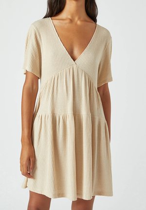 Femme portant une robe beige côtelée à manches courtes avec un décolleté en V profond et une jupe évasée à volants, sur un fond uni.