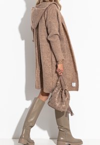 Langer beiger Strickcardigan mit Kapuze, gerippten Ärmeln und einem Etikett. Kombiniert mit hellbraunen knielangen Stiefeln und einer strukturierten Bucket Bag.