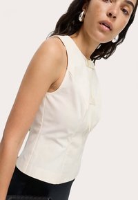 Top sin mangas ajustado en tela color crema claro con una textura suave, con un botón decorativo en la parte delantera y costuras limpias.