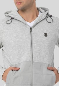 Graue Zip-Hoodie mit strukturiertem Oberteil, glatten unteren Paneelen, Seitentaschen und einem kleinen Logo auf der Brust. Verstellbare Kordelzugkapuze.