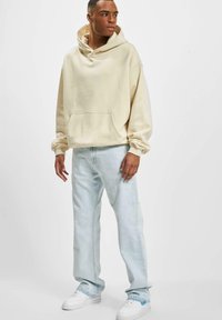 Cremefarvet hoodie med en kængurulomme, parret med lyseblå, løstsiddende jeans og hvide sneakers. Glat tekstur overalt.
