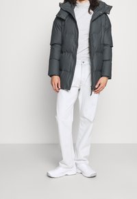 Chaqueta acolchada gris oscuro con capucha, que cuenta con un cierre de cremallera y botón a presión. Lleva puesta una blusa de cuello alto gris claro, combinada con jeans blancos y zapatillas.