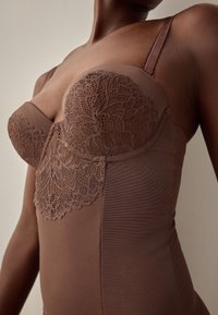 Gros plan d'une personne portant un bustier en dentelle marron avec des bonnets moulés et des bretelles réglables, sur un fond neutre.
