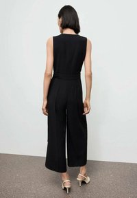 Zwarte mouwloze jumpsuit met een riem in de taille, wijd uitlopende broekspijpen, gladde textuur en minimale details, gedragen met beige hakken.
