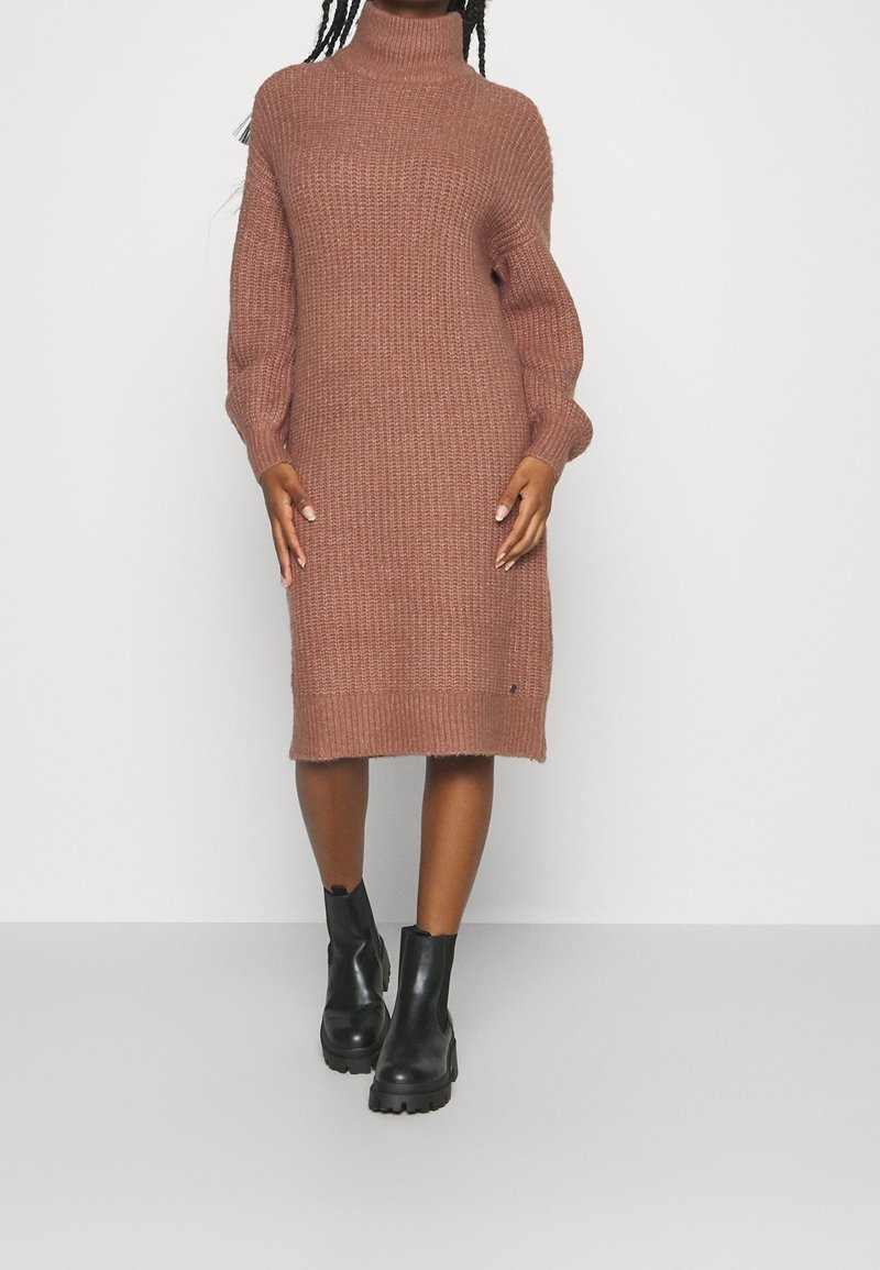 Femme portant une robe marron tricotée épaisse, longueur genou, à manches longues et col montant, avec des bottines noires épaisses, debout devant un fond blanc.