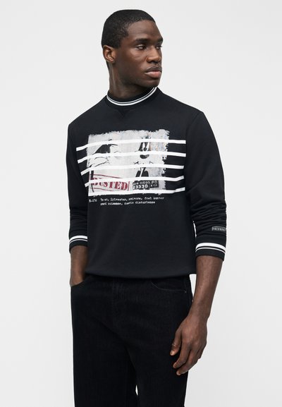 Homme portant un sweat-shirt noir avec des détails à rayures blanches et une impression graphique d'une photo d'identité judiciaire avec du texte, debout les mains dans les poches, regardant vers la droite.