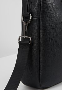 Coin de sac en cuir noir texturé avec fermoir pivotant en métal fixé à une bandoulière noire tissée et réglable.