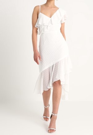 Vestido midi blanco sin mangas con volantes y lunares texturizados, combinado con tacones altos plateados con tiras en el tobillo.