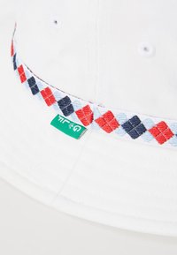 White bucket hat with a patterned woven band featuring red, blue, and black diamonds. Includes a small green tag with branding.