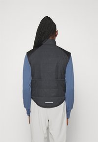 Sort polstret vest med høj krave, der har en buet linning og en vandret hvid reflekterende stribe på bagsiden. Bæres over en blå langærmet top.