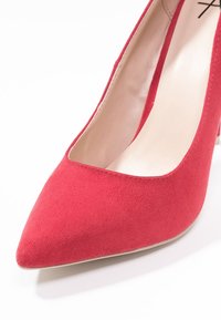 Rode suède pump met een spits toelopende neus, een gladde beige binnenkant en een hak van gemiddelde hoogte. Geen zichtbare versieringen of metalen details.