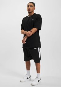 T-shirt oversize noir avec logo blanc, associé à un short noir avec des rayures sur les côtés. Des baskets blanches et des chaussettes côtelées complètent la tenue.