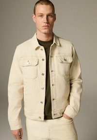 Beige denimjakke med to brystlommer, sølvknapper og krave. Båret over en mørkegrøn t-shirt og parret med lyse bukser.