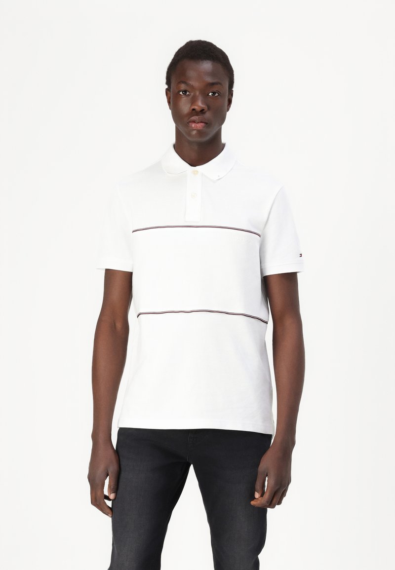 Hvid polo shirt med korte ærmer, der har to vandrette maroon striber. Lavet af tekstureret bomuld med krave og knaplister.