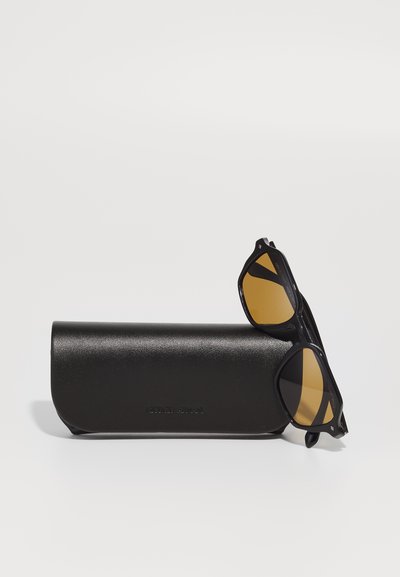 Ochelari de soare negri cu lentile galbene, având un cadru unghiular, stau pe un toc rigid, elegant, de culoare neagră, cu o finisare texturată.