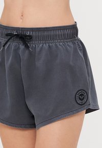 Shorts sportivi grigi con una cintura elastica, lacci neri, tasche laterali e una patch rotonda con logo sulla parte inferiore della gamba sinistra.