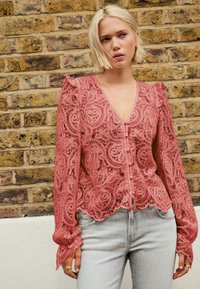 Čipkasta bluza v prahu rožnata barvi z zapletenimi vzorci, dolgimi rokavi in nabranimi poudarki. Gumbi spredaj in rob z valovitimi robovi.
