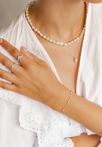 Deux colliers de perles superposés, une chaîne en argent avec un pendentif en perle, une bague en argent aux motifs floraux, et un bracelet délicat en perles.