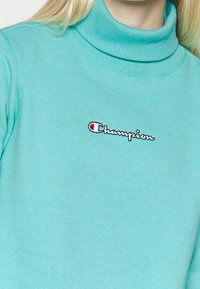 Felpa turchese con colletto alto, realizzata in tessuto morbido. Presenta il logo "Champion" ricamato in bianco e rosso.