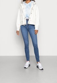 Chaqueta ligera blanca con botones de presión sobre una sudadera con capucha de tie-dye azul cielo, combinada con jeans ajustados azules y zapatillas blancas con detalles.