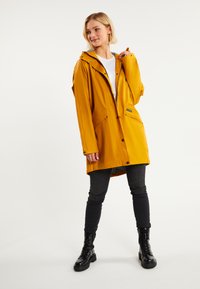 Giacca impermeabile gialla con cappuccio, bottoni frontali, due tasche laterali e fodera grigia. Indossata sopra jeans neri aderenti e stivali neri.