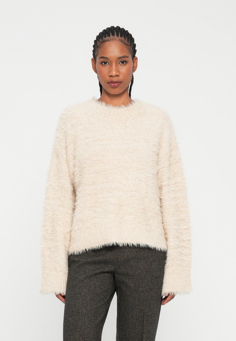 Maglione beige morbido con collo rotondo, maniche lunghe e un aspetto soffice e testurizzato. Combinato con pantaloni grigi a fantasia.