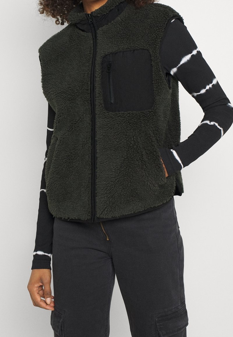 Henkilö, jolla on päällään tummanvihreä fleece-takki, jossa on vetoketjullinen rintatasku, musta-valkoinen kirjontamayttopaita ja mustat cargo-housut.