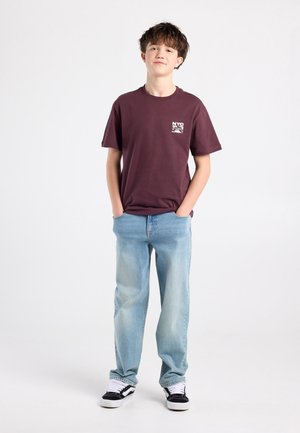 Donkerrode korte mouwen t-shirt met "NYC SK8" grafisch ontwerp, gecombineerd met lichtblauwe jeans met rechte pijpen en zwarte sneakers met witte accenten.