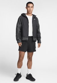 Svart vadderad jacka med huva, bärs över en vit strukturerad skjorta. Stylad med svarta shorts, vita strumpor och svarta sneakers.