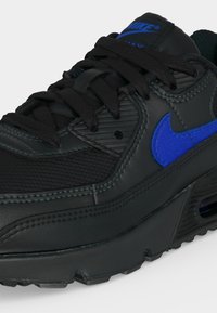 Μαύρο αθλητικό παπούτσι Nike Air Max με μπλε λογότυπο swoosh, μαύρα κορδόνια και υφασμάτινες λεπτομέρειες στην πλευρά και στη σόλα.