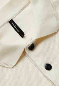 Krämfärgad polo skjorta med texturerad tyg, med en ribbad krage, tre svarta knappstängningar och en svart etikett nära kragen.