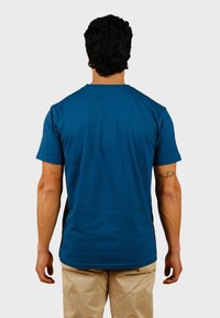 Camiseta de algodón azul con cuello redondo y mangas cortas, mostrada desde atrás. Presenta una textura suave y un pequeño parche con logo en el cuello.