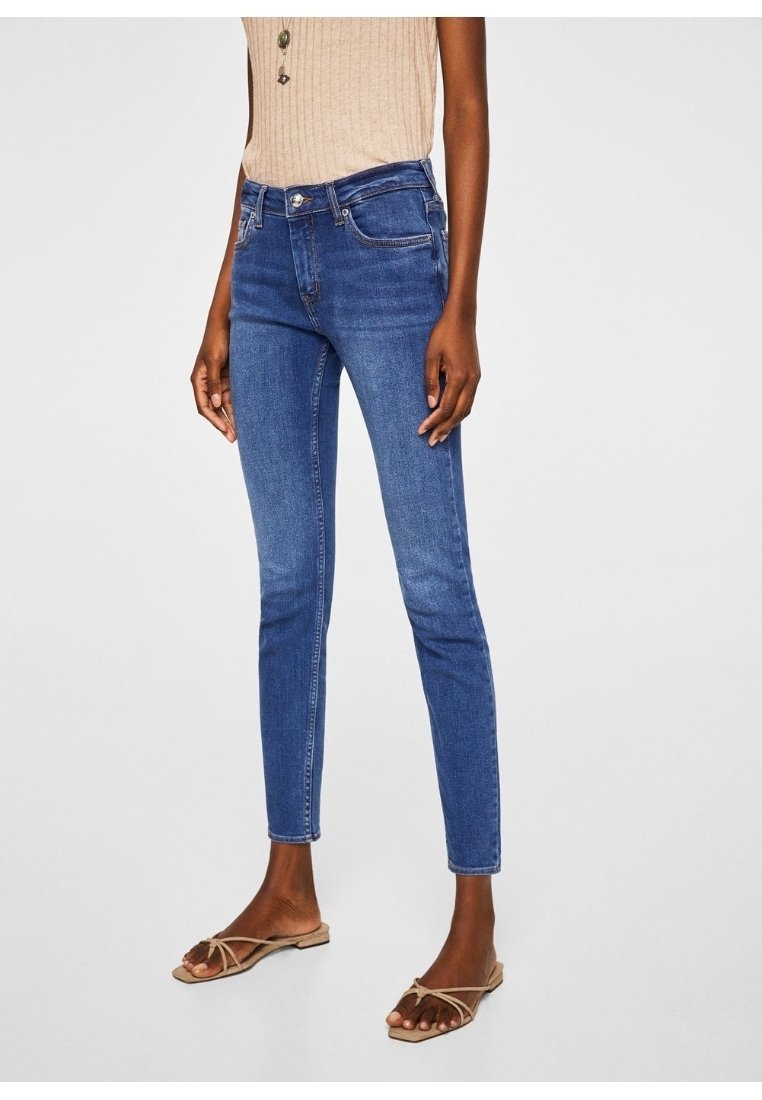 Femme portant un jean slim bleu, un top beige côtelé sans manches et des sandales plates beiges à brides, debout devant un fond clair uni.