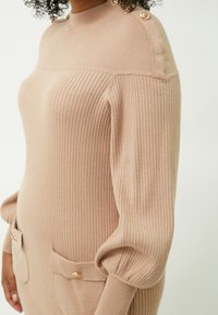Lys beige ribbet sweaterkjole med høj krave, guldknapdetaljer på skuldrene og to forlommer med knaplukninger.
