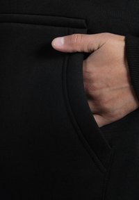 Hand teilweise in der vorderen Tasche eines schwarzen Kleidungsstücks, wobei der Daumen außen und die Finger innerhalb der Tasche sichtbar sind.