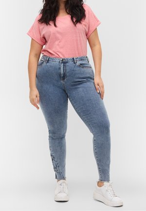 Femme portant un haut rose à manches courtes, un jean skinny bleu taille haute avec des nœuds sur les côtés près des chevilles, et des baskets blanches, debout sur fond blanc.