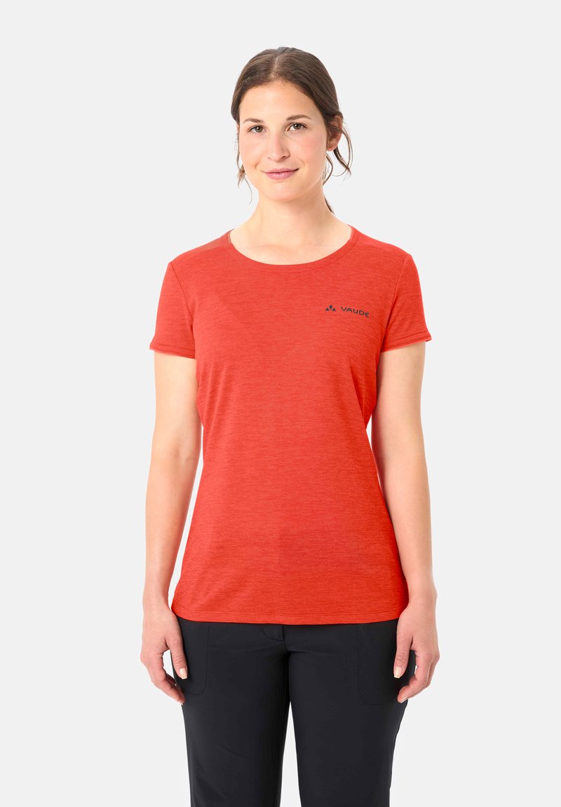 Donna con capelli castani indossa una t-shirt arancione a maniche corte Vaude e pantaloni neri, in piedi davanti a uno sfondo chiaro e uniforme.