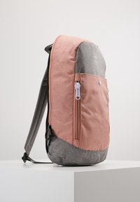 Zaino con materiale superiore rosa e parte inferiore grigia, dotato di una tasca frontale con zipper e dettagli in metallo bianco. Texture complessiva liscia.