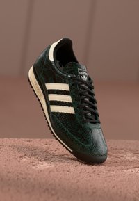 Sneaker sa zelenim uzorkom zmije s krem trakama i crnim vezicama. Ima teksturiranu površinu i kontrastnu bijelu podlogu.