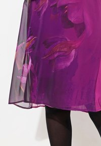 Violetti mekko kukkakuviolla, jossa on suuret terälehdet. Läpinäkyvä ylikerros pehmeällä pinnalla, helma kaartuu lievästi. Mustat sukkahousut näkyvissä.