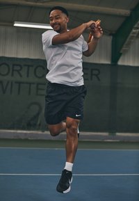 Jugador de tenis con una camiseta blanca ajustada y pantalones cortos negros, sosteniendo una raqueta, realizando un golpe en una cancha azul con un fondo verde.