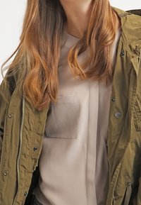 Largo cabello rojo sobre una camiseta beige con bolsillo en el pecho y una chaqueta verde oliva con cremallera y botones, torso superior visible.