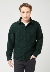 Giovane con capelli chiari che indossa una camicia a maniche lunghe verde scuro a motivo sopra una maglietta bianca, in piedi con una mano in tasca contro uno sfondo neutro.