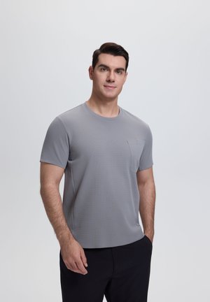 Mężczyzna stoi twarzą do przodu, ubrany w szarą koszulkę z krótkim rękawem z kieszenią na piersi oraz czarne spodnie, na tle jasnego, jednolitego tła.