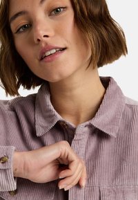 Lichtpaarse corduroy shirt met een traditionele kraag, zichtbare knoopdetails en een geribde textuur, open gedragen bij de hals.