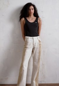 Camisola preta e jeans de cintura alta na cor creme com detalhes em marrom desbotado, em pé contra uma parede clara texturizada.
