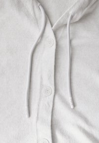 Vêtement à capuche en polaire blanche avec un devant à boutons, présentant une surface texturée, un cordon blanc et quatre boutons ronds.