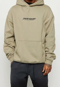 Sudadera de color caqui claro hecha de tela suave, con un bolsillo frontal y el texto "PROBLEMAS DE CONFIANZA" en negro en el pecho. Corte holgado con puños de canalé.