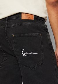 Black denim jeans featuring a brown leather patch with embossed logo and white embroidered detail on the back pocket. Classic five-pocket design.